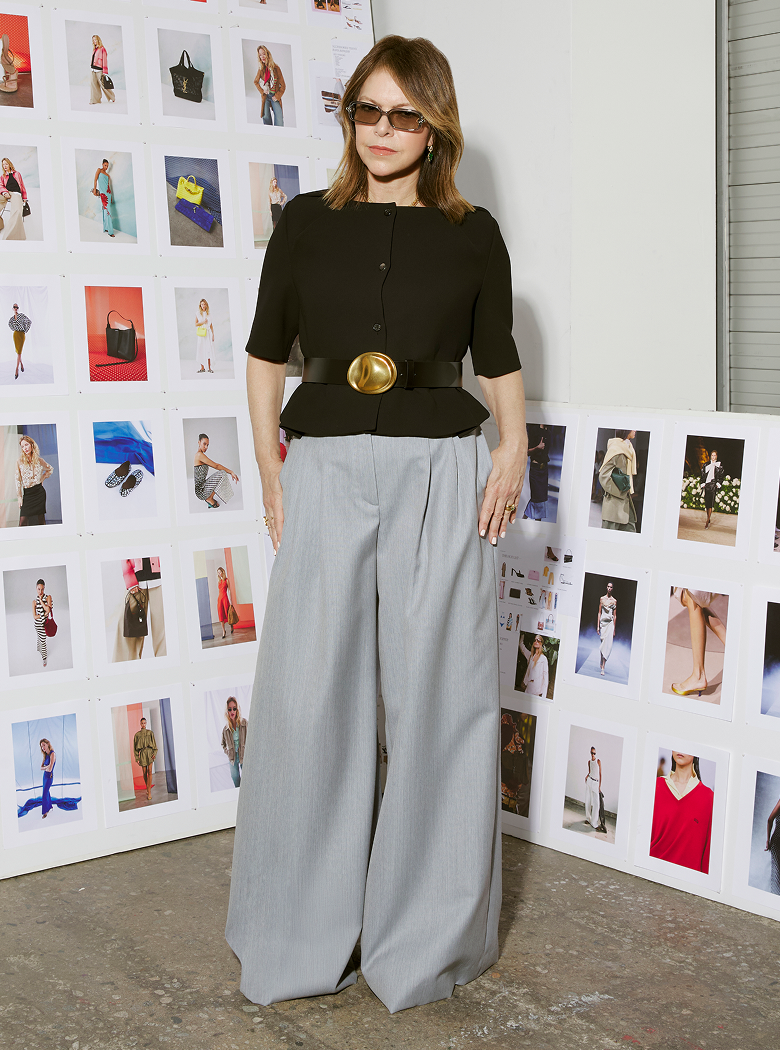 Woman in black top and gray pants standing in front of a wall with multiple small photographs.