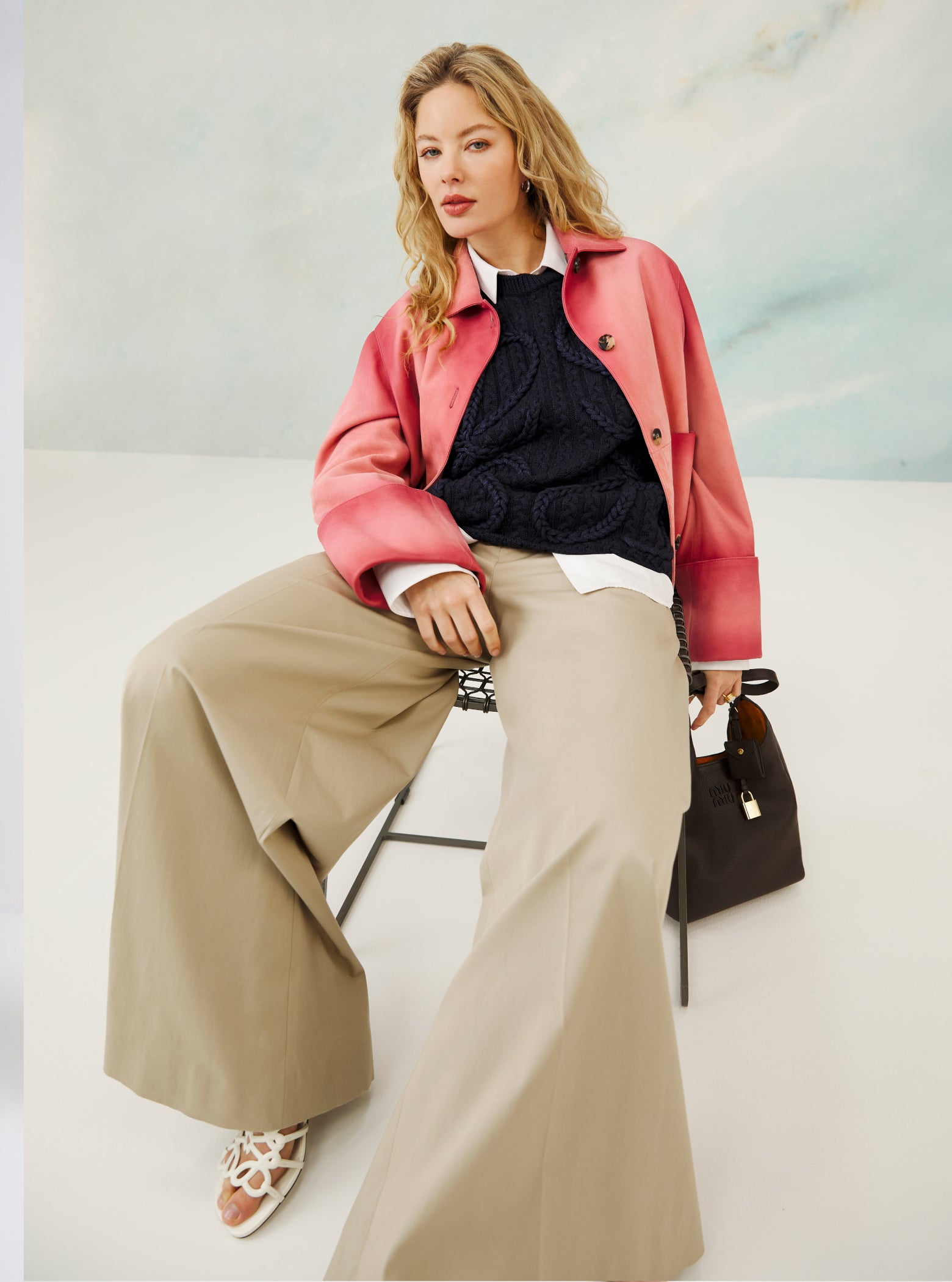 Woman sitting on a chair wearing a pink coat, black sweater, beige pants, and holding a handbag.
