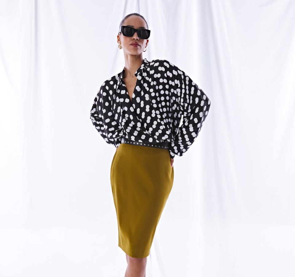 Woman wearing a black and white polka dot blouse with a yellow pencil skirt against a white curtain background.