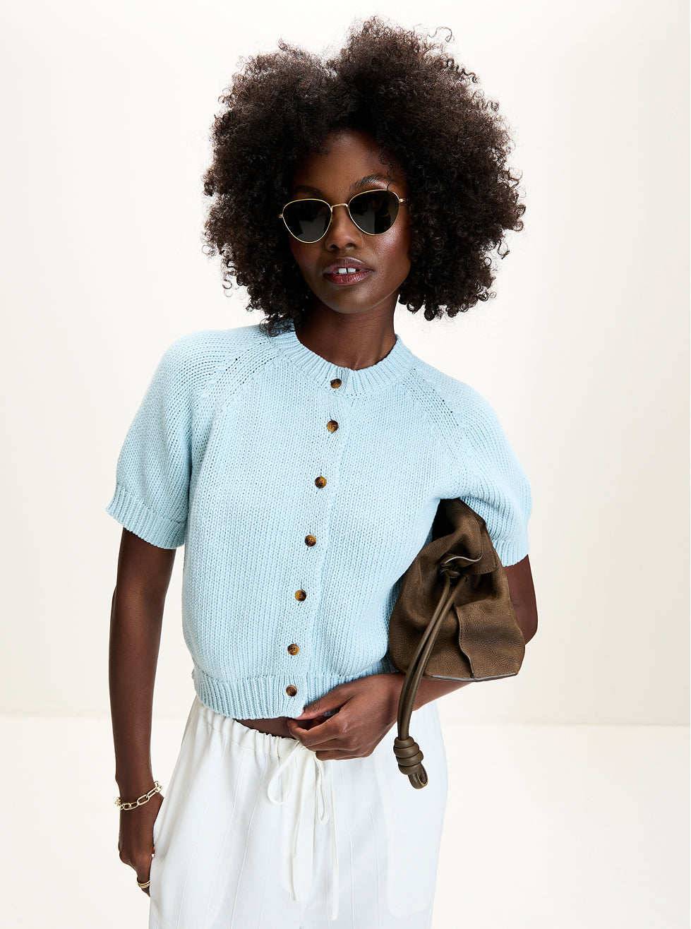 Woman wearing a light blue cardigan with brown buttons, white pants, and holding a brown leather bag against a white background.