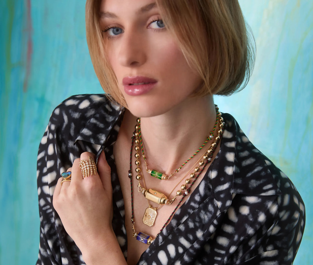 Lady wearing necklaces and rings on her left hand, in a black and white top, with a blue background