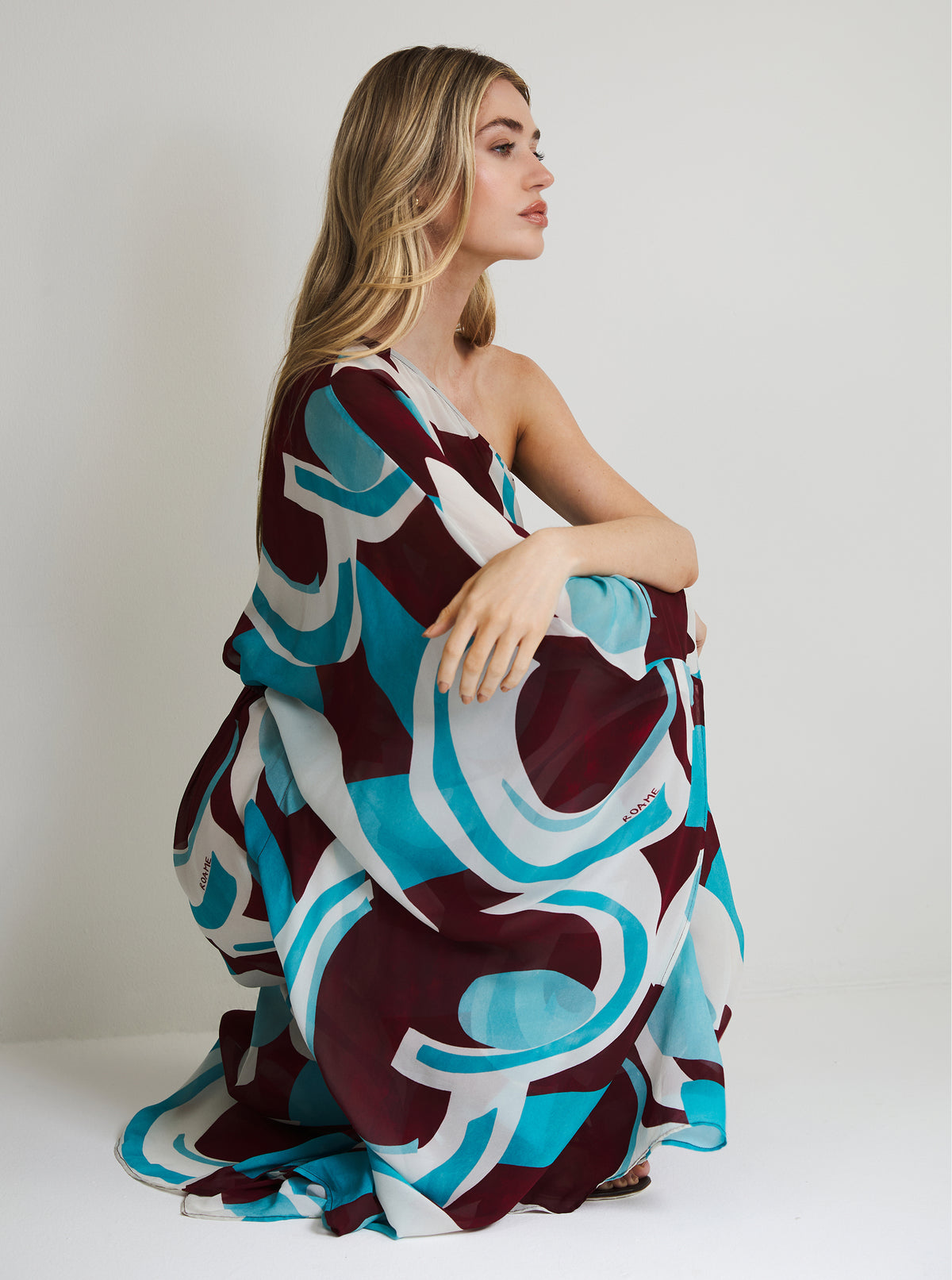 A woman wearing a flowing one-shoulder dress with an abstract wavy pattern in teal, white, and deep burgundy, posed against a plain light gray studio background.