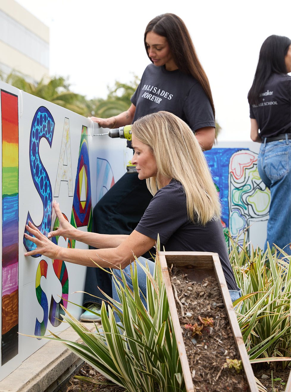 Three women are collaborating outdoors to paint a large colorful mural on a white wall. The woman in the foreground is applying paint to a large letter, while another woman uses a drill on a vertical section.