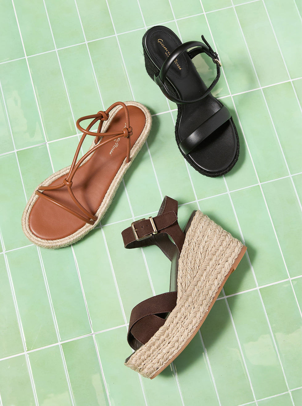 Three women's espadrille-style sandals arranged on a light green tiled background: a black strappy sandal, a tan lace-up flat sandal, and a dark brown wedge sandal with a braided jute platform.