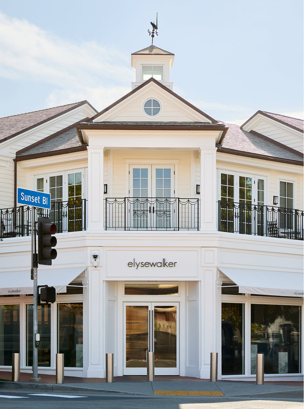 A white two-story retail building with a cupola, black metal railings, large glass windows, and the brand name 'elysewaker' centered above the entrance. A blue 'Sunset Bl' street sign is visible on the left, with a street intersection and sidewalk in the foreground.