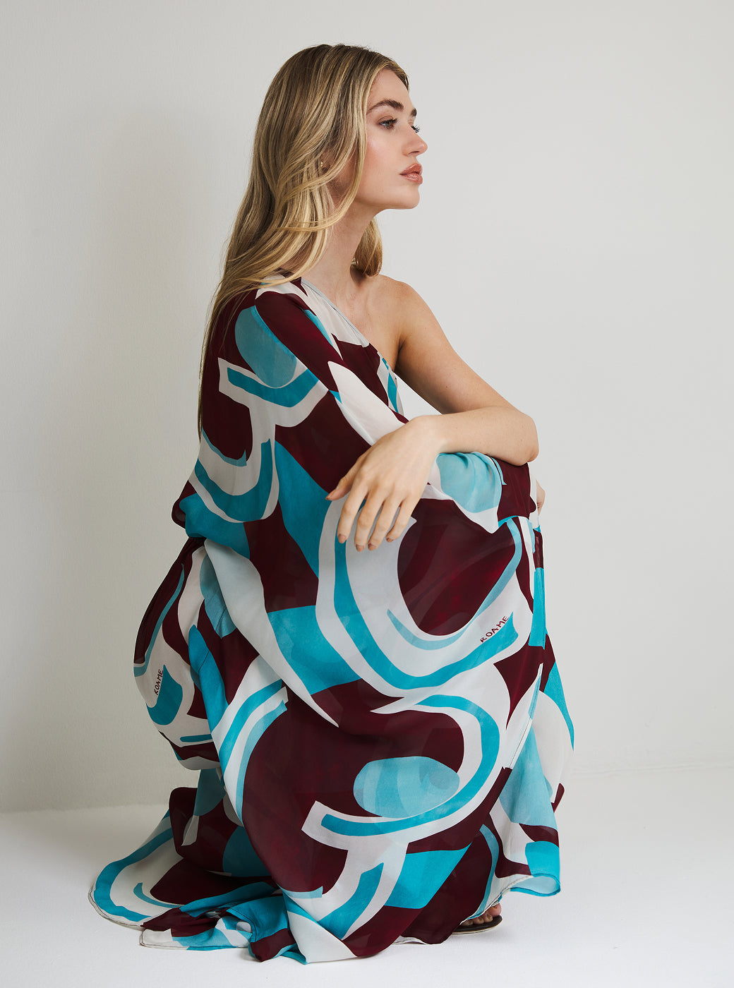 A woman in profile wearing a flowing abstract print wrap or dress in teal, burgundy, white, and gray, posed against a plain light gray studio background.