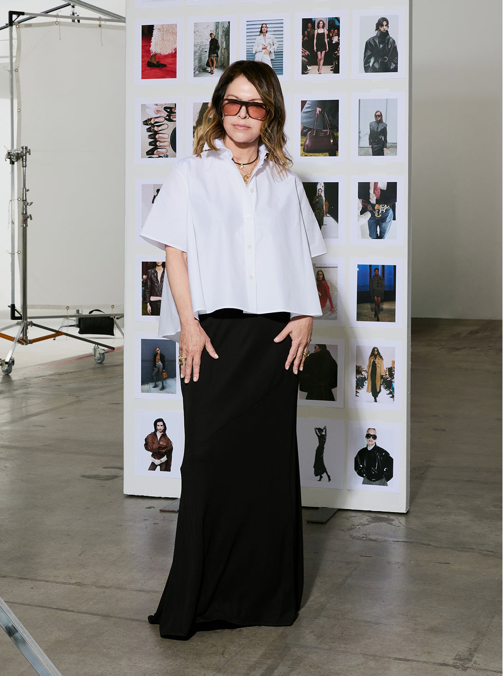 A woman stands in a studio setting wearing a white short-sleeve button-up shirt and a long black skirt. She is positioned in front of a white display board filled with fashion photos.
