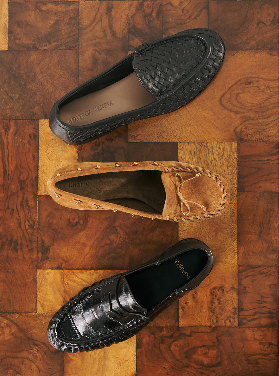 Three Lothario Vettura loafers arranged diagonally on a polished wood block floor. The top shoe is black woven leather, the middle is tan suede with whipstitching, and the bottom is black croc-embossed leather.