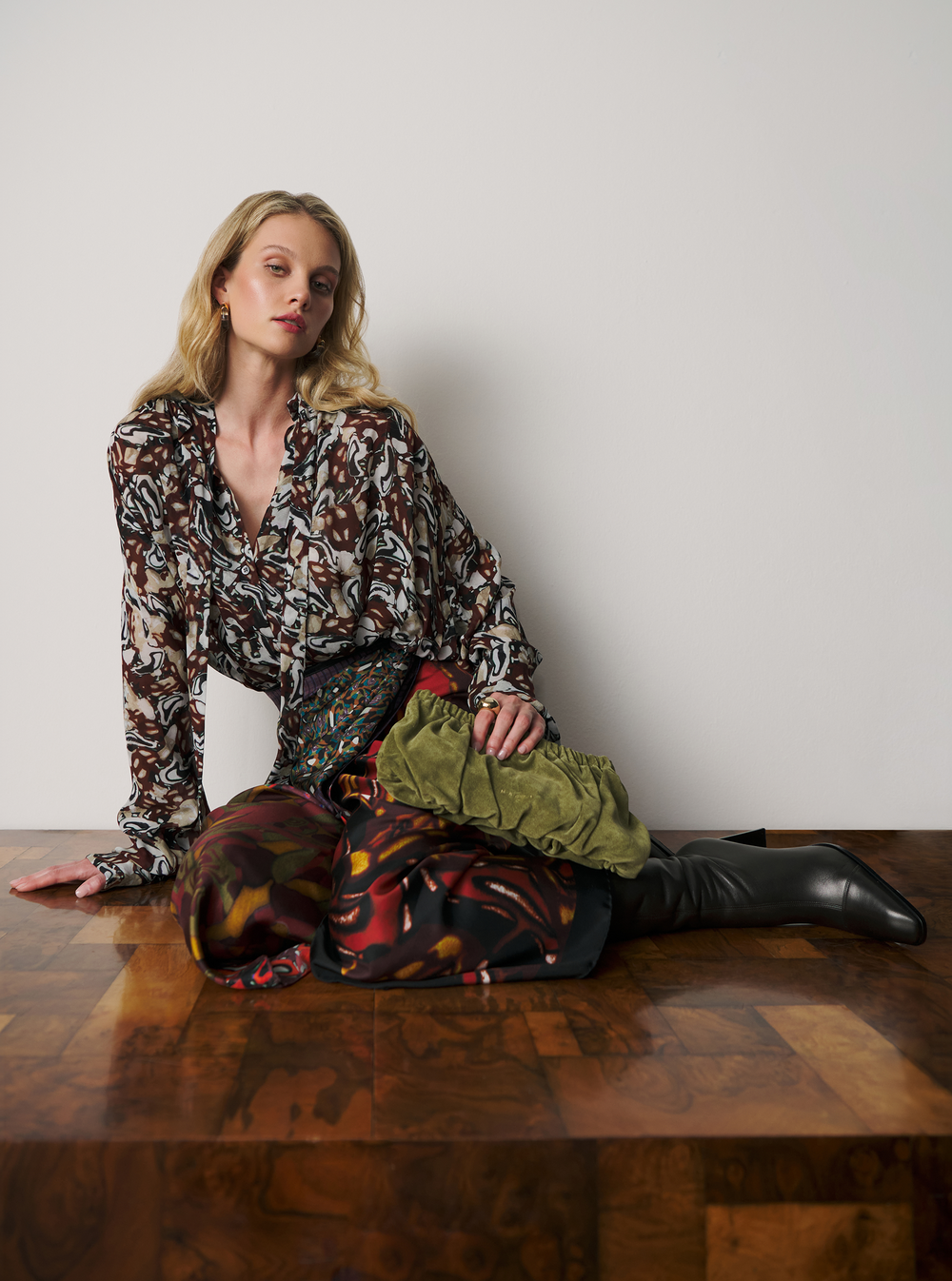 A woman models a patterned long-sleeve blouse and matching patterned skirt while sitting on a wooden table against a plain light gray wall.