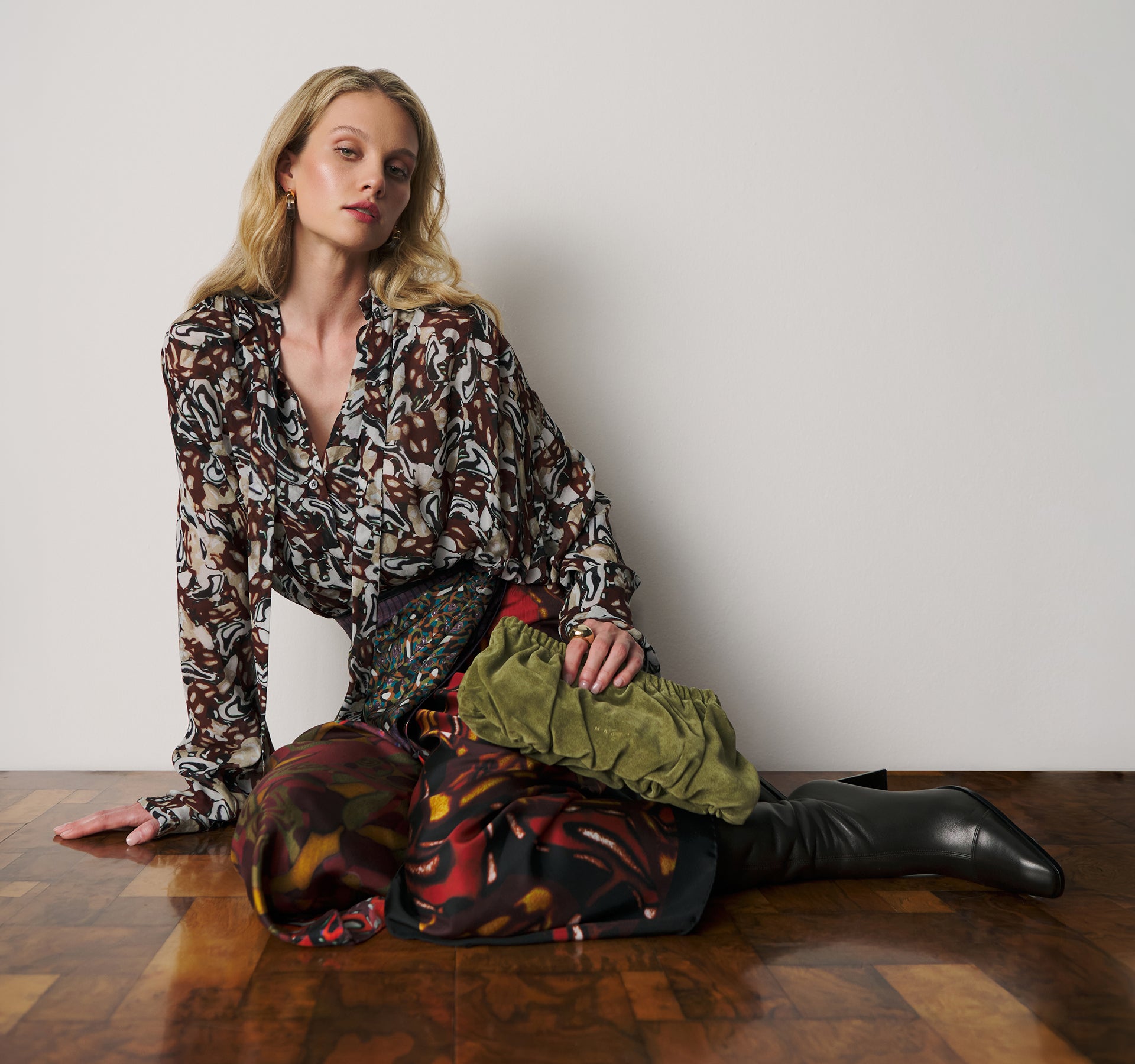 A model sits on a wooden floor against a plain light gray wall, wearing a long-sleeve patterned blouse, a colorful patterned skirt, and black knee-high boots. A folded olive green fabric item rests on her lap.