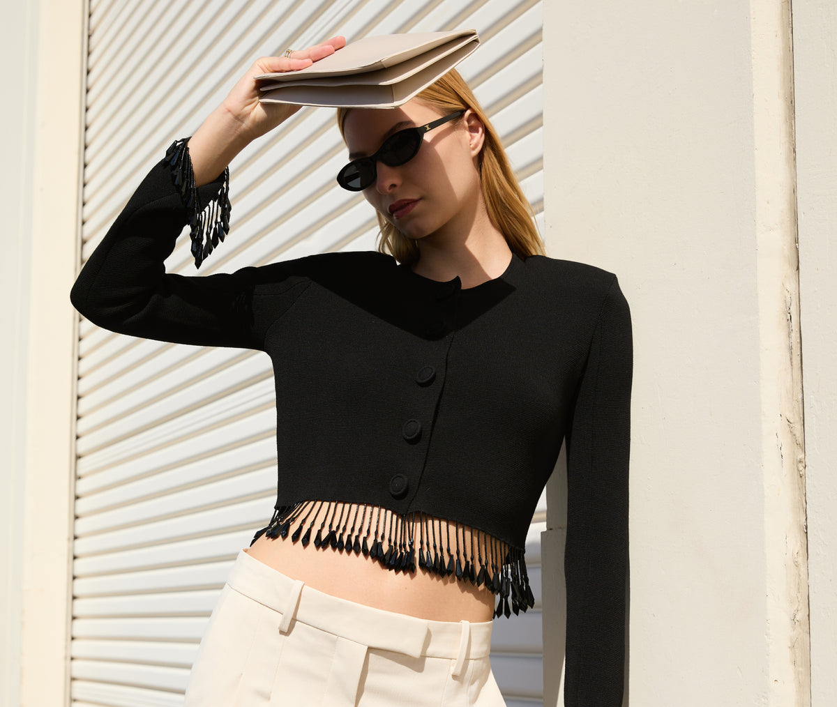 A woman wearing a black long-sleeve cropped cardigan with fringe trim and black sunglasses, paired with cream pants, posing against a white slatted wall while holding a beige clutch.