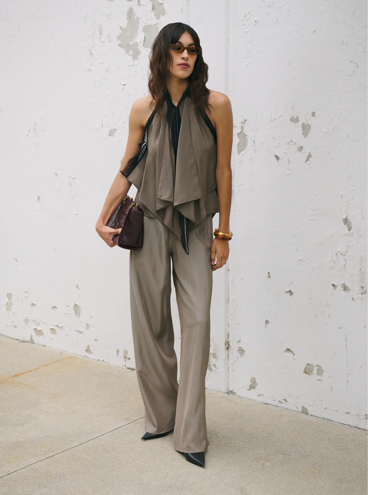A woman stands against a weathered white wall wearing a taupe-beige sleeveless draped top and matching wide-leg trousers, with black pointed shoes and a dark brown handbag.