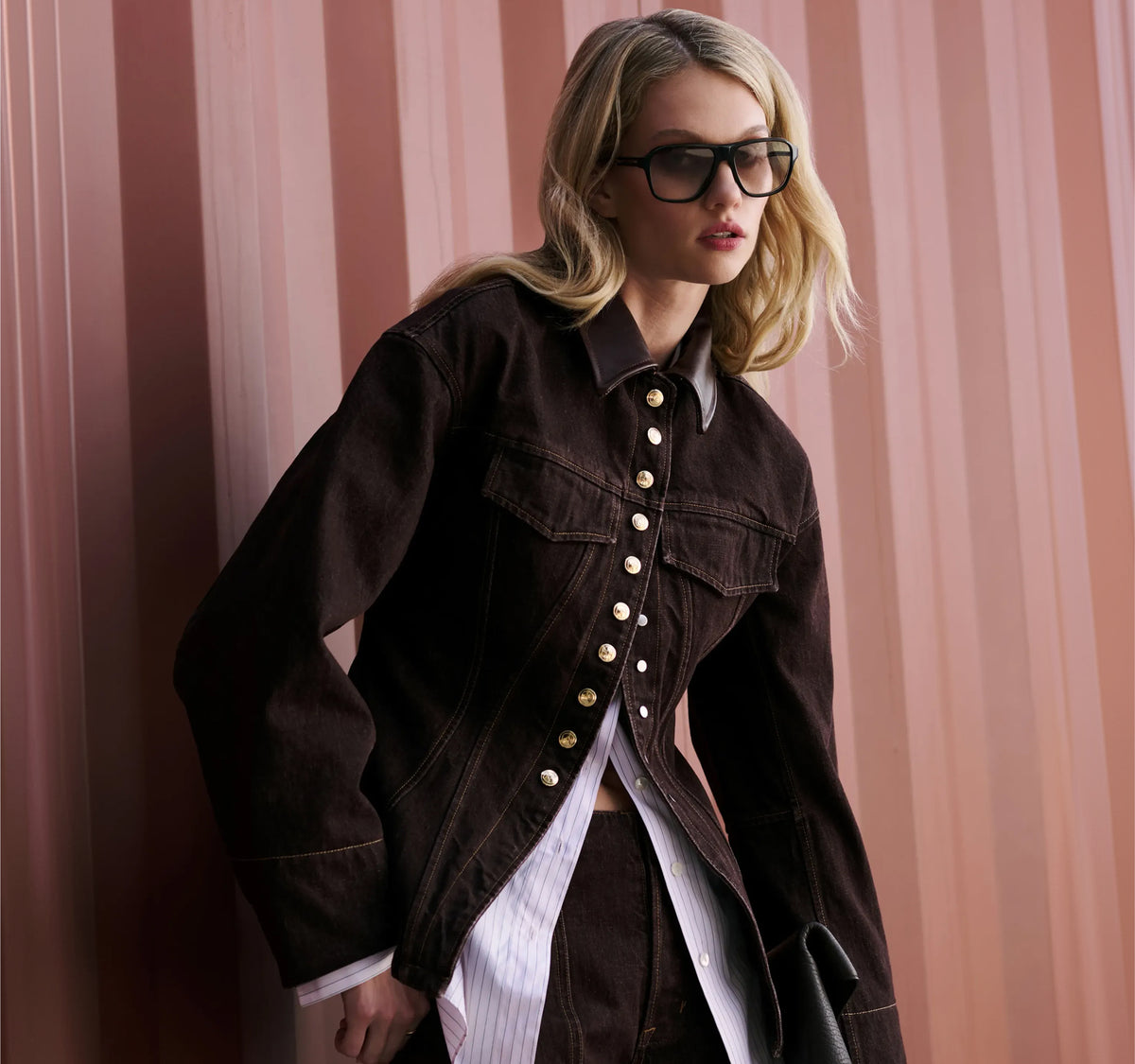 A woman wearing a dark brown denim jacket with gold-tone buttons, styled over a white striped shirt, posing against a pink corrugated metal wall.