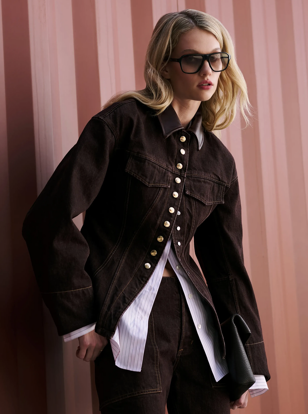 A blonde model wearing a dark brown cropped denim jacket with gold-tone buttons over a white striped shirt, paired with matching dark brown jeans and black sunglasses, posed against a pink striped background.