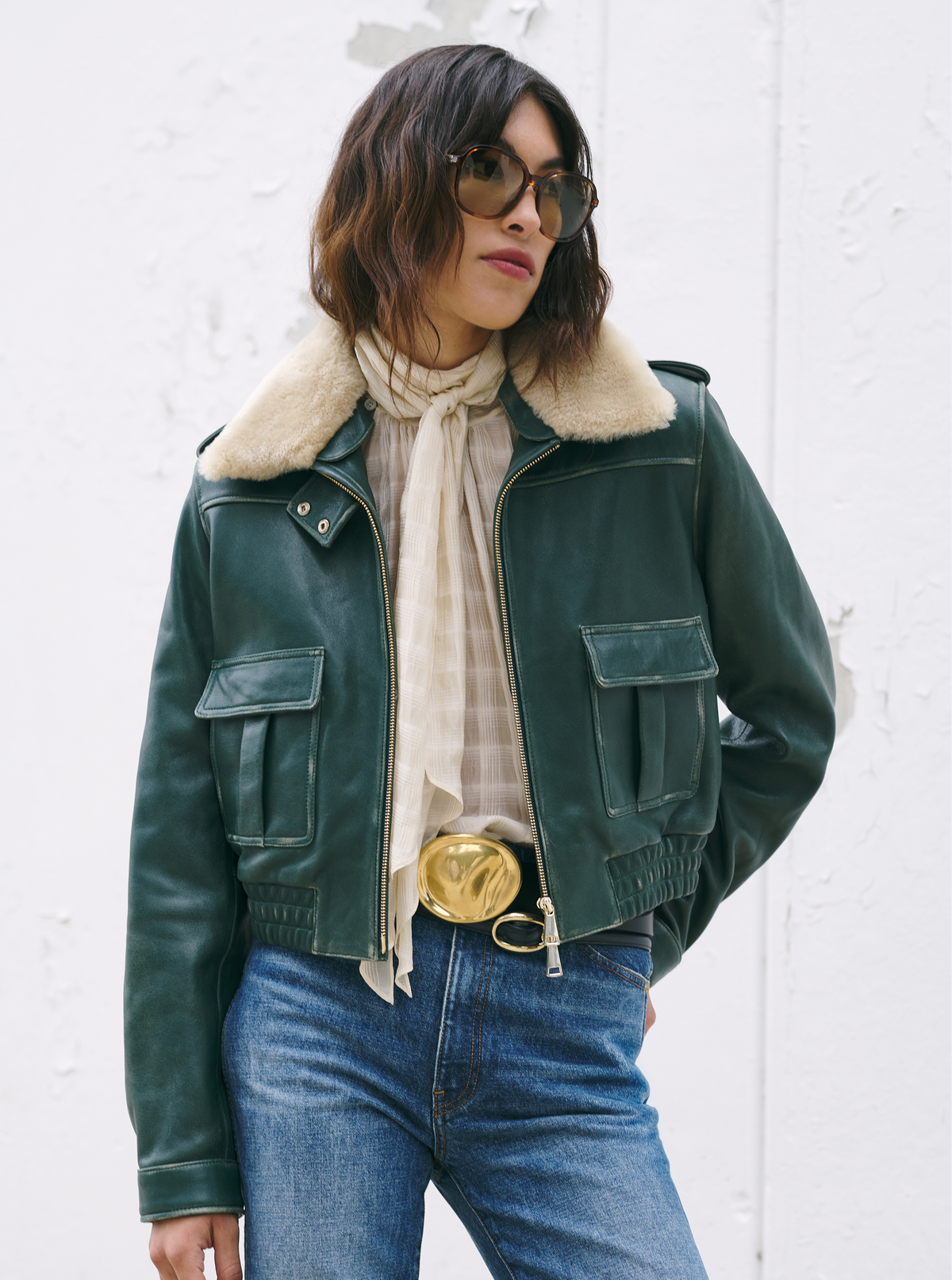 Model wearing a green jacket, a white blouse, a black belt with a gold buckle, and blue jeans standing in front of a white background.