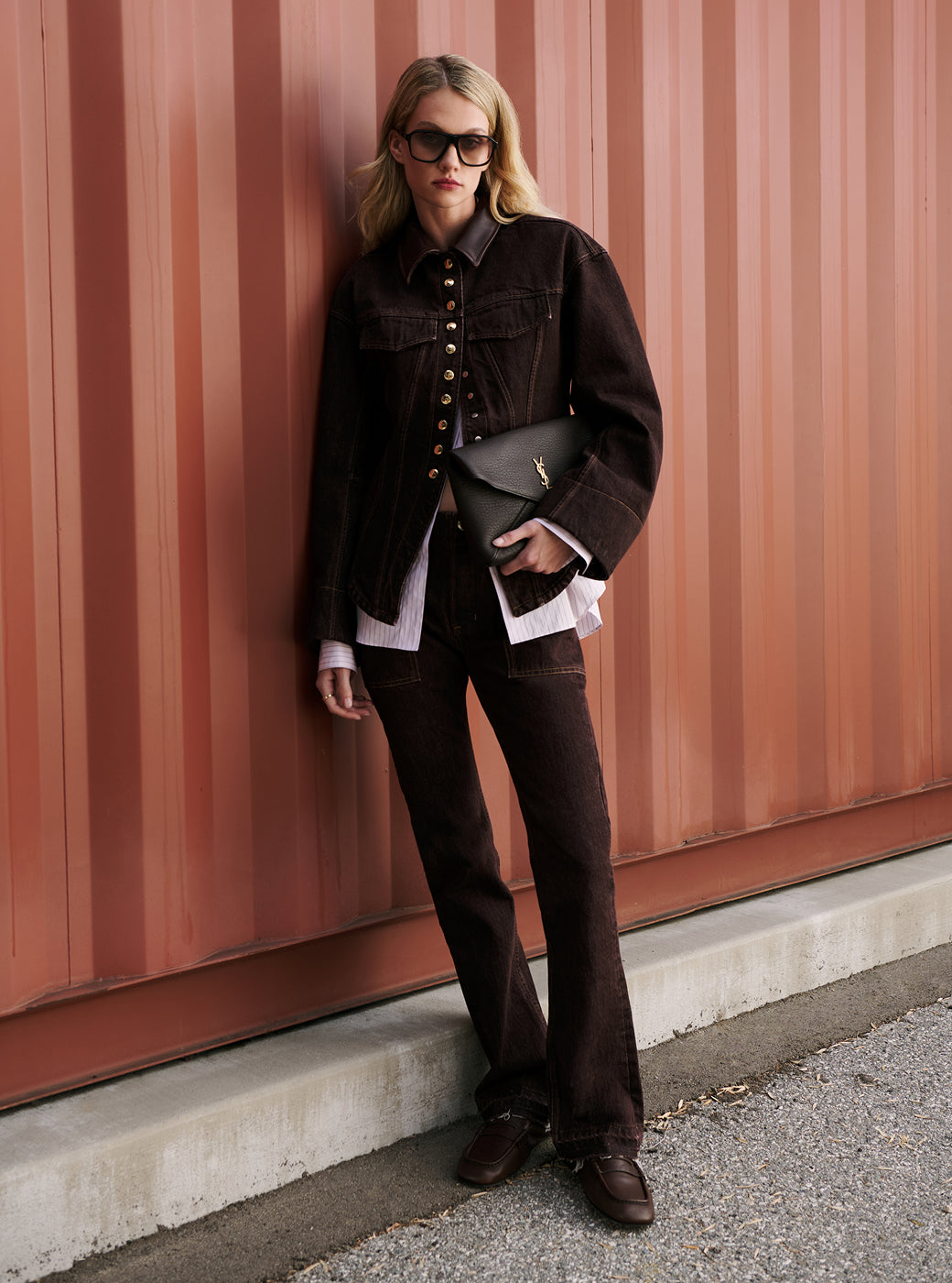 Model standing in front of a pink background wearing a brown denim set, brown sunglasses, brown flat loafers, holding a YSL black clutch purse.