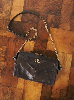 A black leather shoulder bag with a chevron pattern, gold-tone chain strap, and a prominent interlocking V logo on the front, displayed on a polished wooden parquet floor.