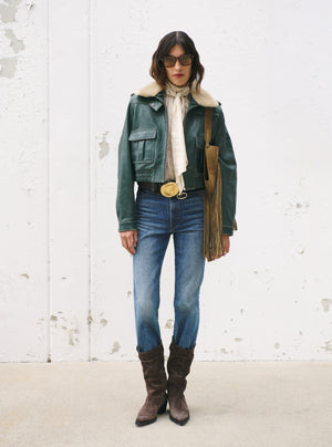 A woman stands against a distressed white wall wearing a dark green leather jacket with a cream shearling-style collar, blue jeans, brown knee-high boots, and carrying a tan fringe shoulder bag.