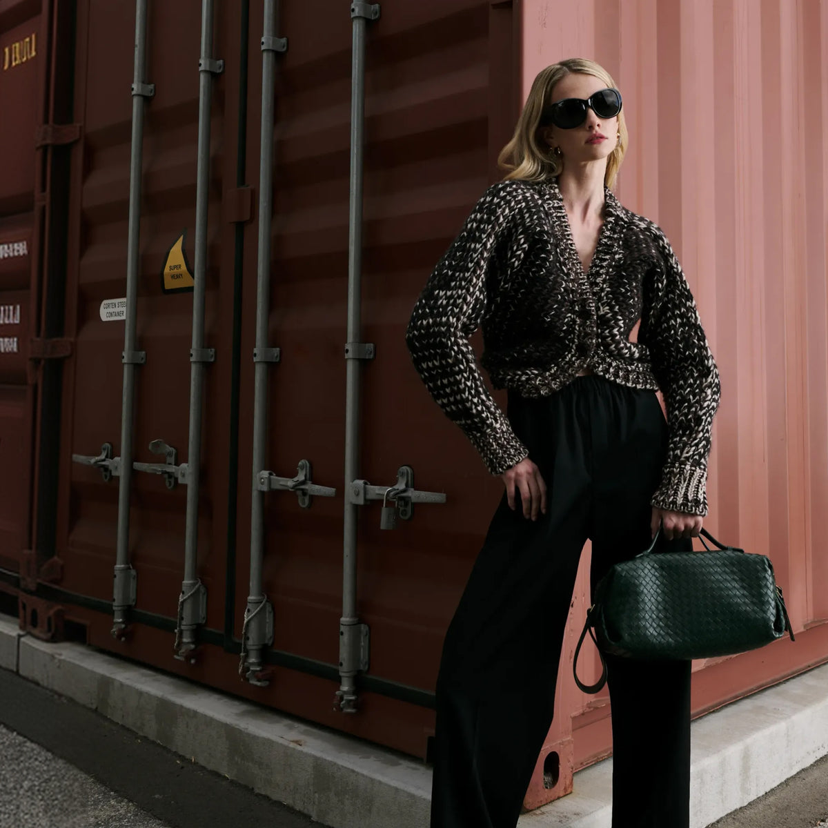 A woman stands in front of a large red-brown shipping container, wearing a black-and-white patterned long-sleeve top, black wide-leg pants, dark sunglasses, and carrying a dark green structured handbag.