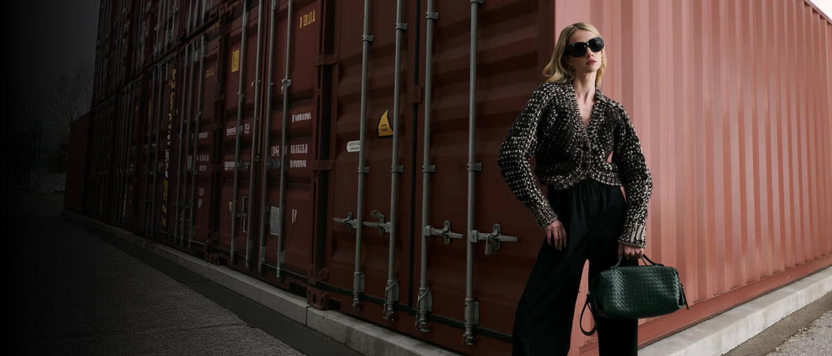 A woman stands in front of a large red-brown shipping container, wearing a black-and-white patterned long-sleeve top, black wide-leg pants, dark sunglasses, and carrying a dark green structured handbag.